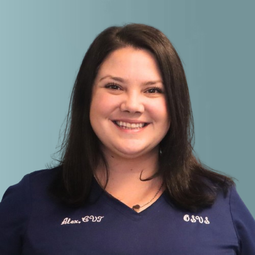 Smiling woman with dark hair wearing a navy blue shirt embroidered with a name and initials, stands against a plain light teal background.