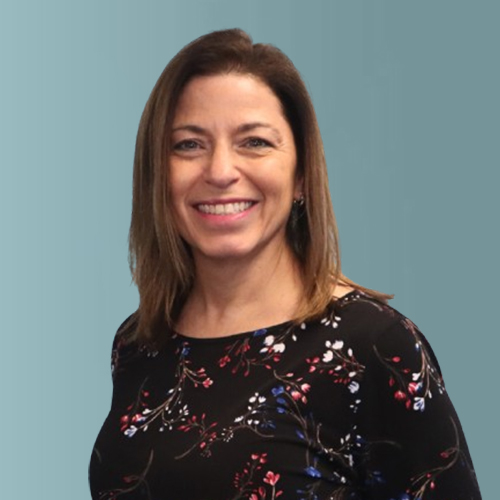 A woman with shoulder-length brown hair is smiling at the camera. She is wearing a black top with a floral pattern. The background is a plain teal color.