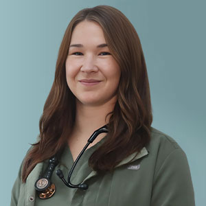 A person with long brown hair and a friendly smile is wearing a dark blue vest over a maroon top, standing against a plain gray background.