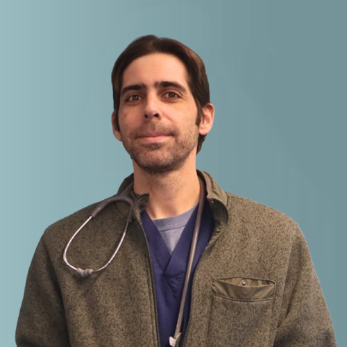 A person with short hair is wearing a stethoscope around their neck, a green fleece jacket, and a blue shirt. The background is a plain teal color.