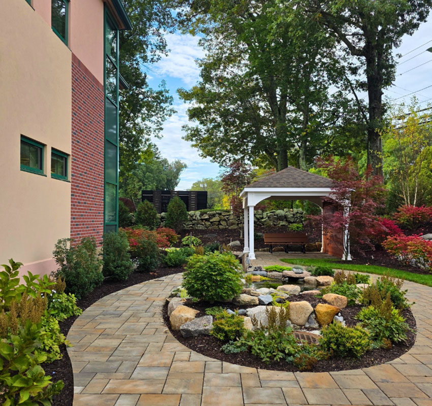 A landscaped garden with a circular stone path, central rock garden, lush plants, and a white gazebo with a swing, next to a brick building under tall trees and a partly cloudy sky.