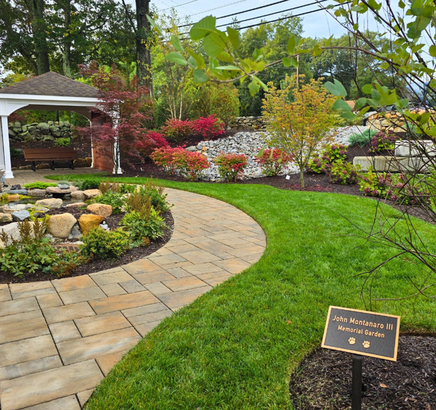 A neatly landscaped garden with a curved stone path, green lawn, various shrubs, and a gazebo. A sign reads, “John Montanaro III Memorial Garden.” Rocks and colorful plants decorate the area.