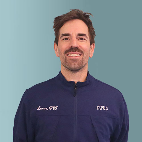 A person with short brown hair and a beard wearing a navy blue jacket with embroidered text. The background is a solid teal color.