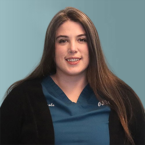 A person with long brown hair is smiling at the camera. They are wearing a blue top and a black cardigan, standing against a solid light blue background.