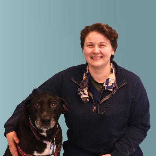 A person wearing a blue sweater and a stethoscope around their neck smiles while sitting next to a black dog with a white muzzle, against a blue-gray background.