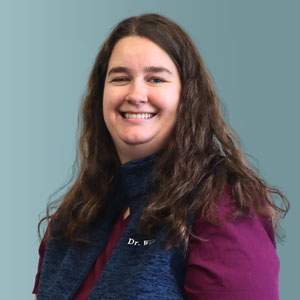 A person with long brown hair and a friendly smile is wearing a dark blue vest over a maroon top, standing against a plain gray background.