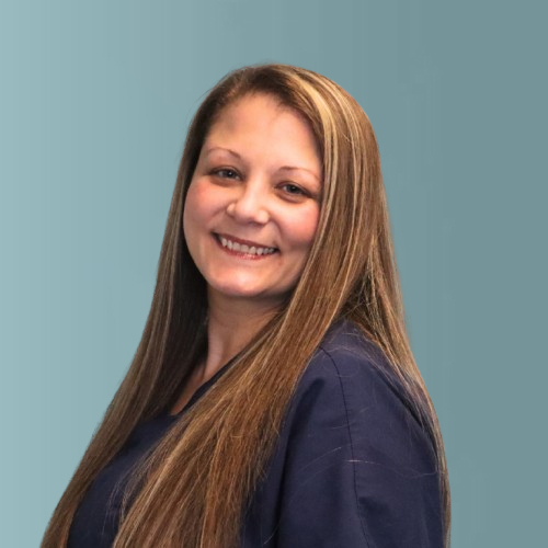A person with long brown hair is smiling at the camera, wearing a navy blue shirt. The background is a soft teal color.