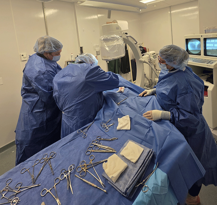 Three medical professionals in blue surgical gowns and masks perform a procedure in a well-lit operating room, surrounded by surgical instruments and equipment. A monitor and sterile tools are visible in the foreground.