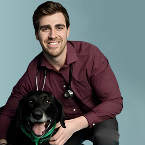 A smiling man in a maroon shirt with a stethoscope around his neck sits next to a happy black dog wearing a green collar, against a plain blue background.