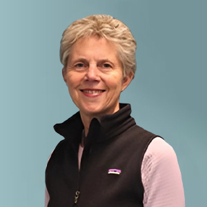 An older woman with short gray hair smiles at the camera. She is wearing a black vest over a light pink long-sleeve shirt. The background is a solid light blue color.