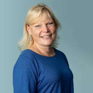 Smiling woman with blonde hair wearing a blue long-sleeve top, standing in front of a plain light blue background.