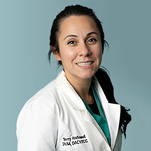 A woman with long dark hair tied back, wearing a white lab coat and green scrubs, smiles in front of a plain light blue background.