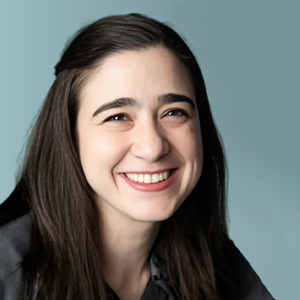 A woman with long dark brown hair and fair skin smiles warmly at the camera. She is wearing a dark top and is posed in front of a plain, light blue background.