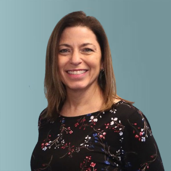 A woman with straight brown hair, wearing a black floral-patterned top, smiles at the camera against a plain teal background.
