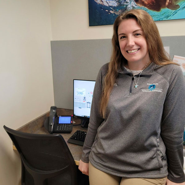 A woman with long hair, wearing a gray logo pullover, is smiling and standing next to an office desk with a computer, phone, and chair. A large photo is on the wall behind her.