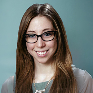 A woman with long brown hair and glasses is smiling at the camera. She is wearing a translucent-sleeved top and a necklace, with a plain teal background behind her.