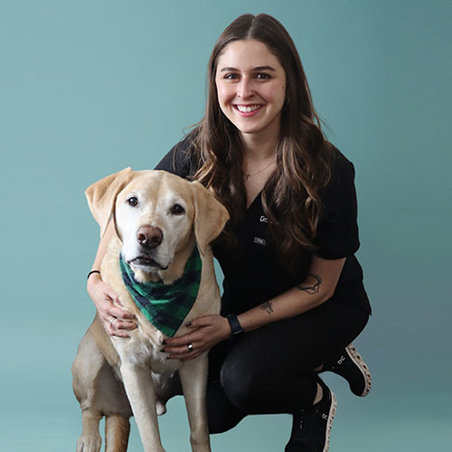 A woman with long brown hair, wearing black clothes, smiles while kneeling next to a yellow Labrador retriever dog wearing a green plaid bandana, against a plain teal background.