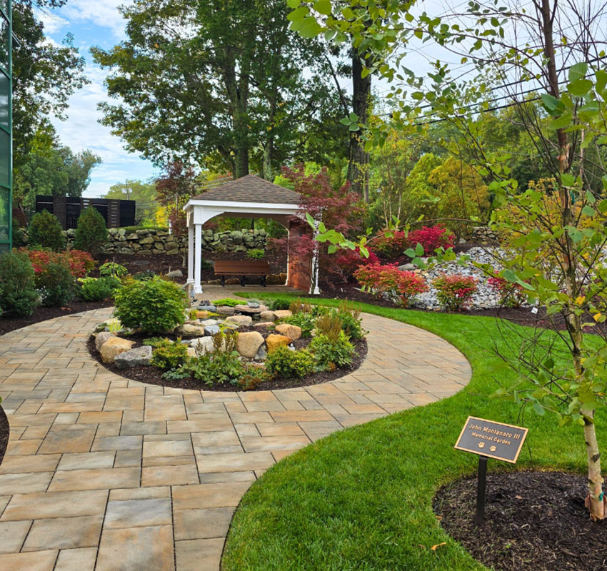 A landscaped garden features a winding stone path, vibrant flower beds, a small pond with rocks, and a white gazebo surrounded by trees and shrubs. A brown sign is staked in the grass near the path.