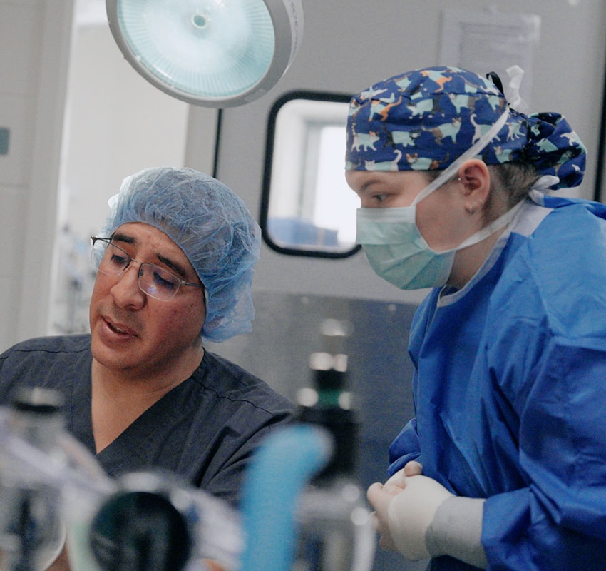Two medical professionals in scrubs, masks, and surgical caps are talking in an operating room. One is seated and looking down while the other stands beside him, listening attentively. Medical equipment is visible in the foreground.