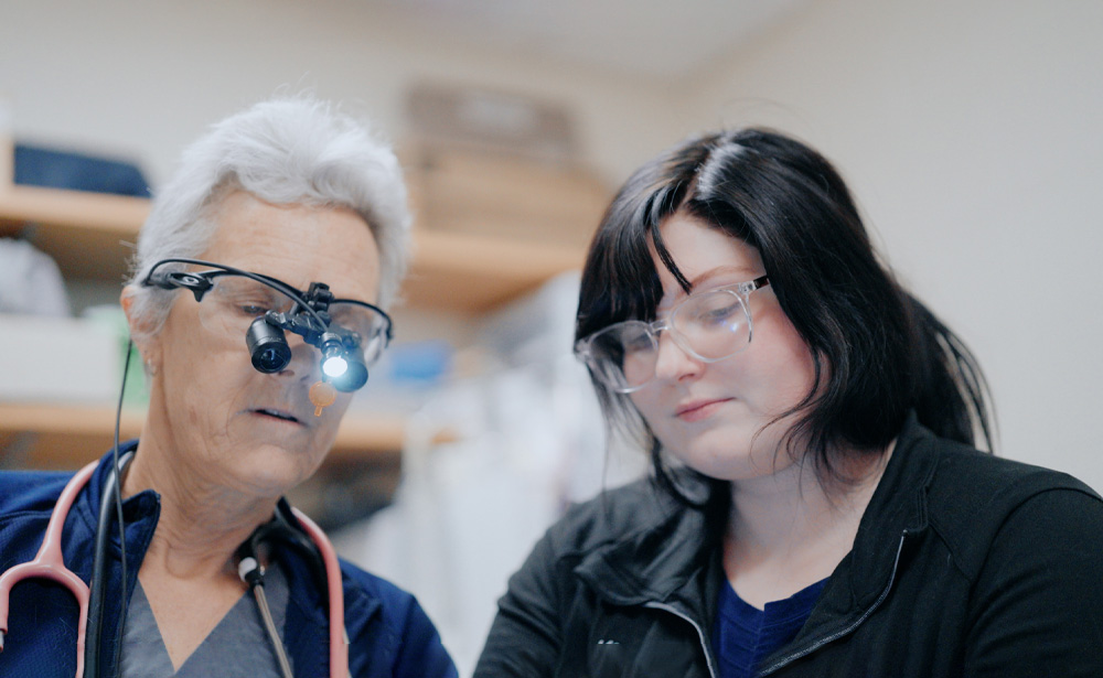 A healthcare professional wearing magnifying glasses with a headlamp and a stethoscope examines something with a young woman, both focused and wearing protective glasses in a medical setting.