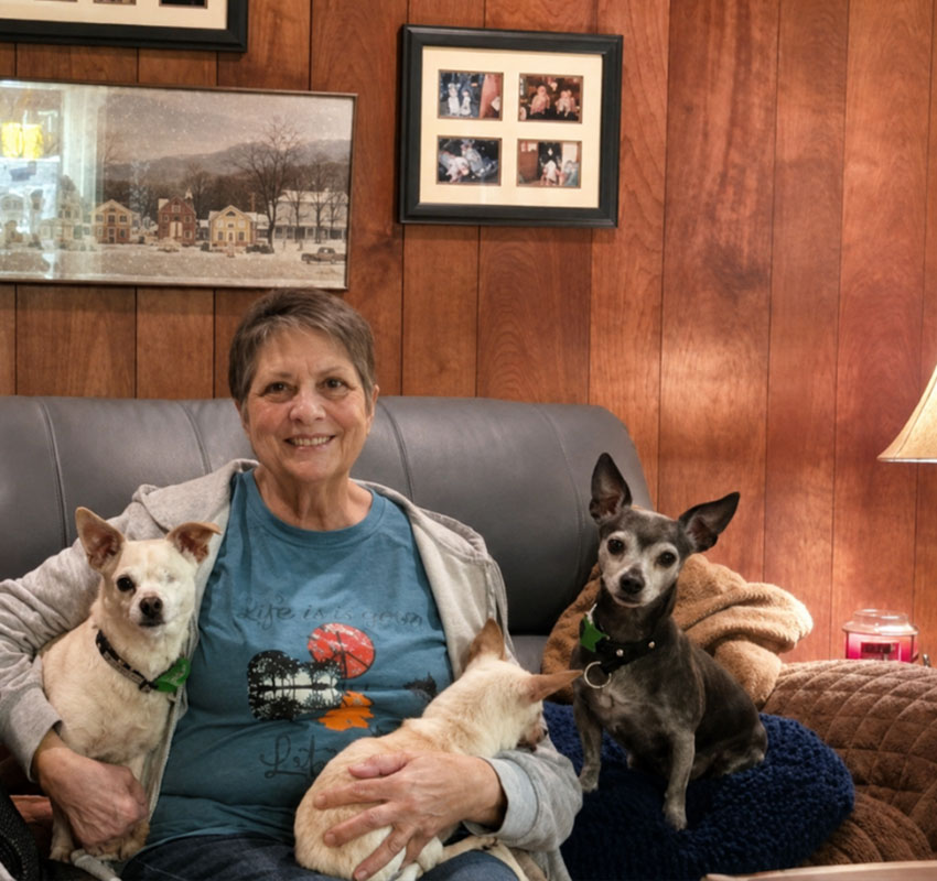 Smiling woman sits on a couch with three small dogs on her lap. The background features wood-paneled walls, framed photos, a lamp, and cozy blankets.